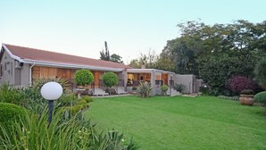 Una casa de dos pisos con techo de tejas rojas, césped cuidado y diversas plantas y arbustos.