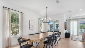 A spacious dining area with a long wooden table and modern chairs, large windows with curtains, and a view of a building outside.