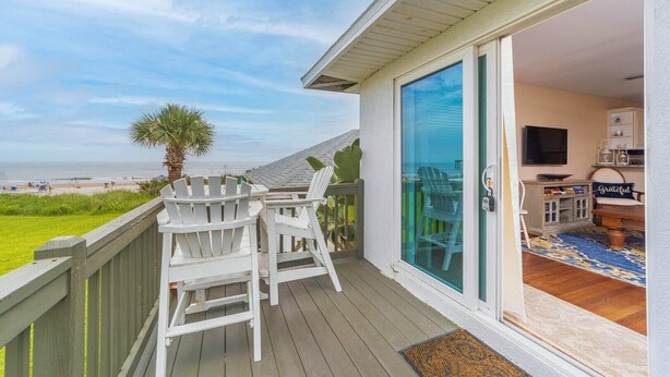 Un balcon avec des fauteuils Adirondack blancs, une porte vitrée donnant sur un intérieur équipé d’un téléviseur et orné d’un coussin sur lequel est inscrit le mot « Grateful », et une vue sur la plage.