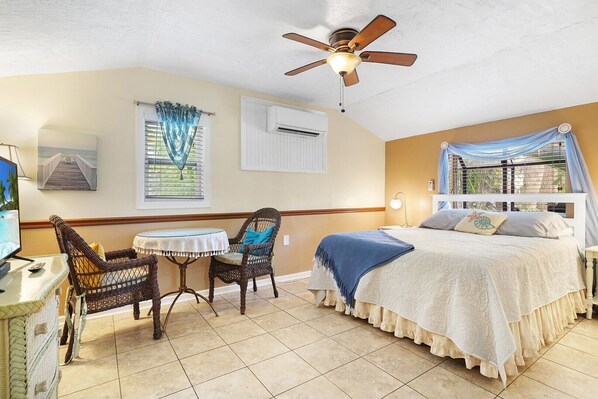 A bedroom with a bed, a ceiling fan, a window with curtains, a chair, a table, and a TV. - 8.5 B Street - Beachside Bungalow (St. Augustine Beach)