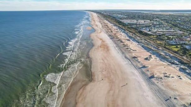 An aerial view of a coastal area with a sandy beach, a clear blue ocean, and a developed urban area with buildings and roads.