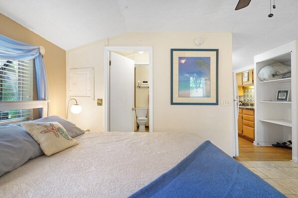 A bedroom with a bed, a nightstand, a bedside table, a window with curtains, a door leading to a bathroom, and a kitchen area visible through an open door.
