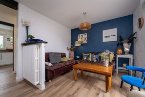 Sala de estar con sofá, mesa de centro y sillas azules. Se observa un radiador, una lámpara colgante y obras de arte en la pared.