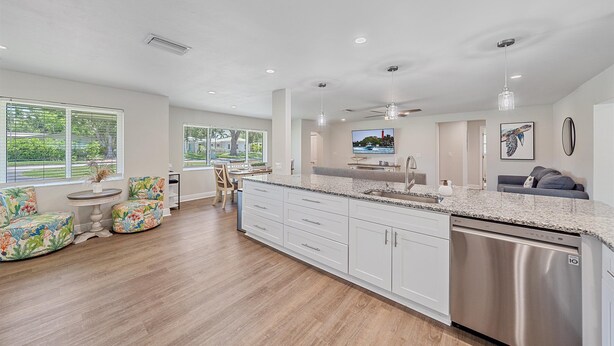 A modern kitchen with white cabinetry, a stainless steel dishwasher, and a granite countertop. There are two chairs with floral upholstery and a small round table.