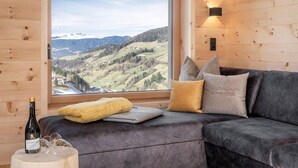A cozy room with a large window offering a scenic view of a valley, a dark grey sofa with cushions, a bottle of wine, and glasses on a small table. - Apartment 'Mittermanting' with Mountain View, Private Terrace and Wi-Fi (Villnoss)