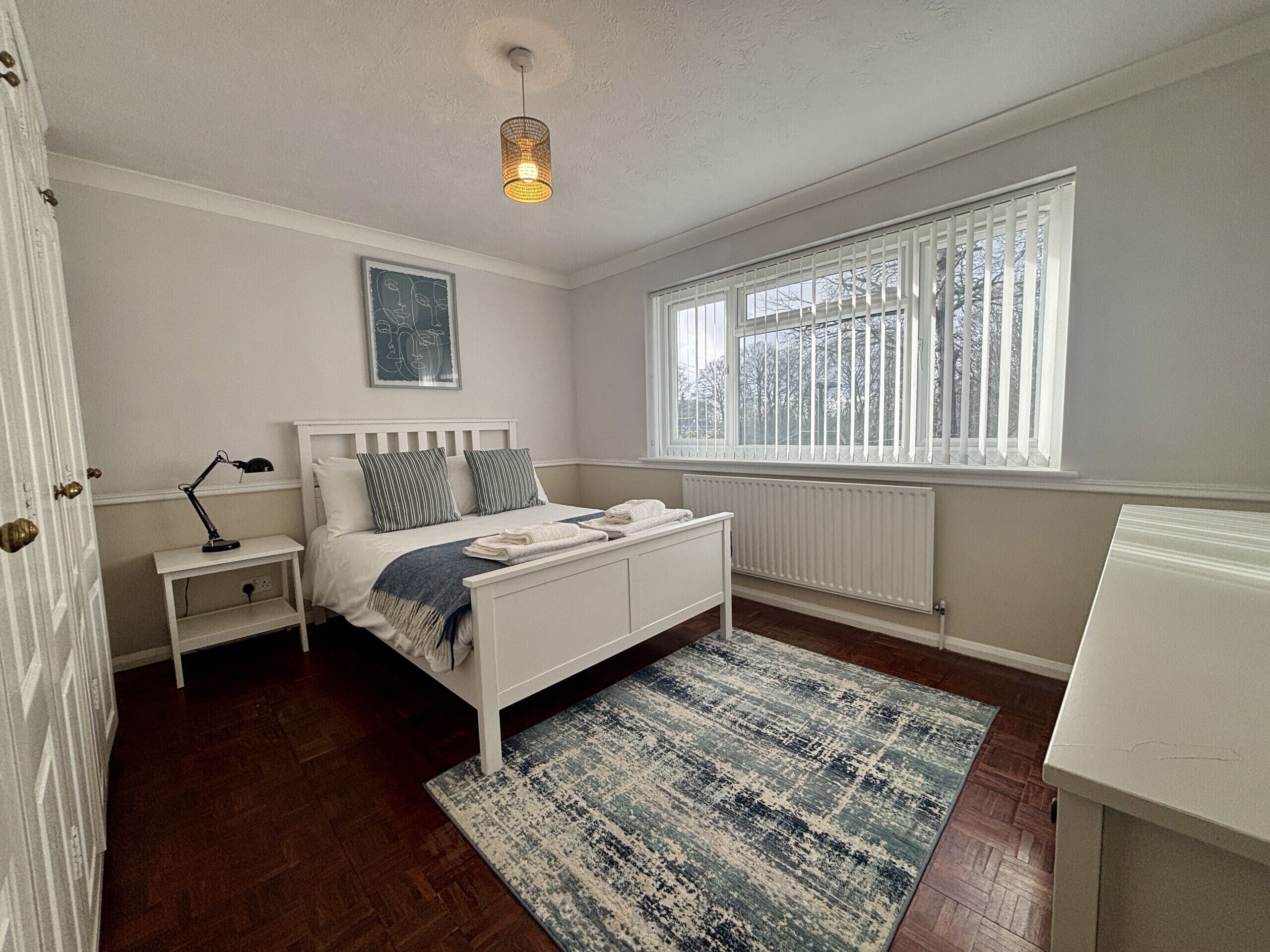 A bedroom with a bed, bedside table, lamp, window, radiator, and a door.