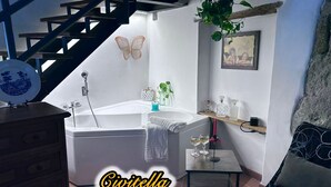 Un baño con bañera, lavamanos y una pequeña mesa con dos vasos. Se ve una escalera al fondo y una planta en la pared.