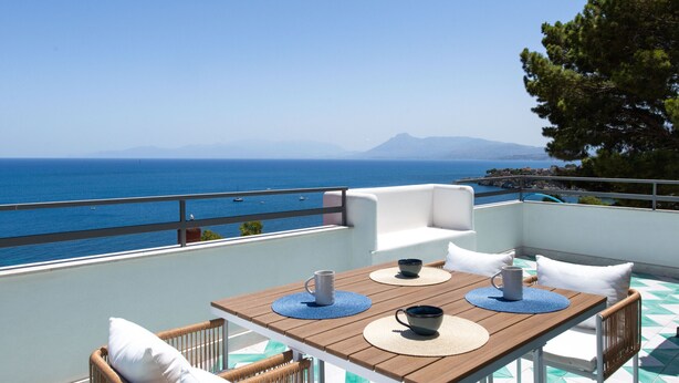 Una terraza con mesa de madera, cojines blancos y sillas de mimbre, con vistas a un mar azul despejado con barcos y montañas a lo lejos.