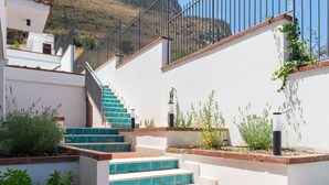 Una escalera con escalones azules que sube a un balcón, rodeada de paredes blancas y follaje.