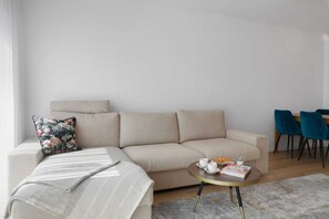 Un salon moderne avec un canapé beige, un coussin à motifs floraux, un plaid rayé et une table basse ronde sur laquelle se trouvent un livre et un verre d’eau.