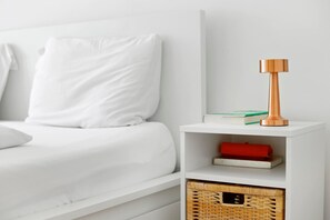 A white bed with a white pillow, a white nightstand with a book and a red item, and a woven basket.
