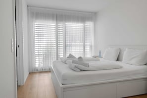 A modern, white-themed bedroom with a large bed, a bedside table, and sheer curtains.