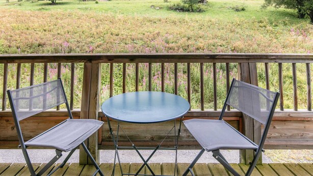 Une terrasse en bois avec deux chaises en métal et une table ronde, donnant sur un champ verdoyant parsemé de fleurs sauvages.