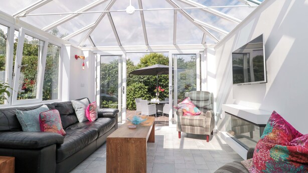 A conservatory with a glass ceiling, a black leather sofa, a wooden coffee table, and a plaid armchair.