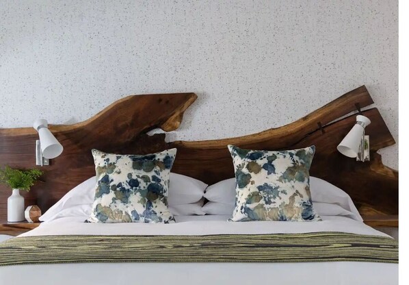 Ein Bett mit Holz-Kopfteil, zwei passende Kissen, ein Nachttisch mit Lampe und eine kleine Pflanze.