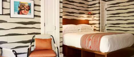 Une chambre avec un mur à rayures zébrées, un lit avec un cadre en bois, une chaise avec des coussins orange et un tableau encadré accroché au mur.