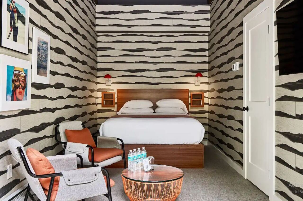 A compact hotel room with a bed, two bedside tables, a chair, a small round table, and a wall with a striped pattern.