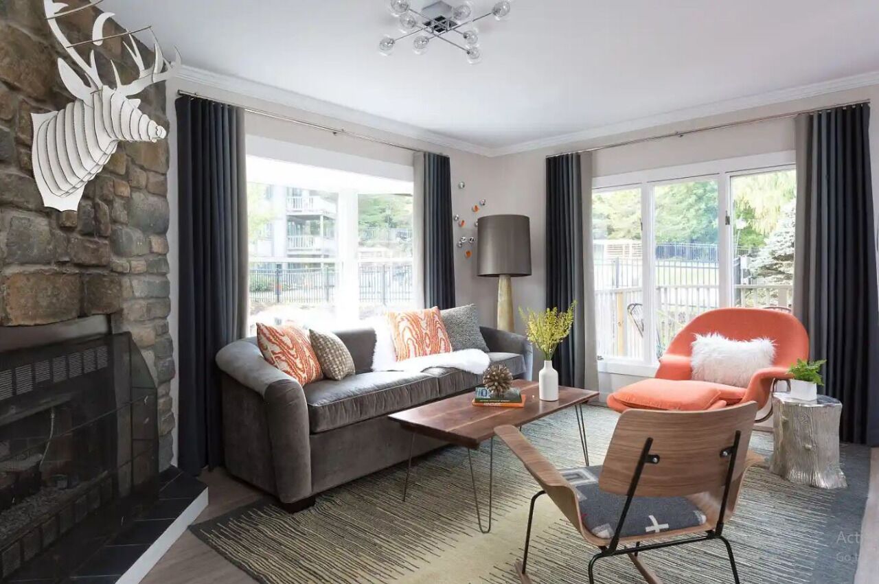 Una sala de estar moderna con chimenea de piedra, un sofá gris, un sillón naranja y un ventanal con cortinas.