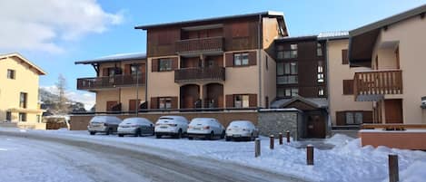 A multi-story building with balconies, a parking area with cars, and a snow-covered ground.