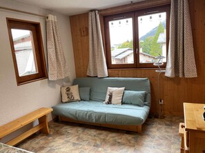 A room with a light blue sofa, two pillows, a wooden bench, a window with curtains, and a view of houses and mountains. - Quiet apartment with balcony and WIFI in Aussois (Aussois)