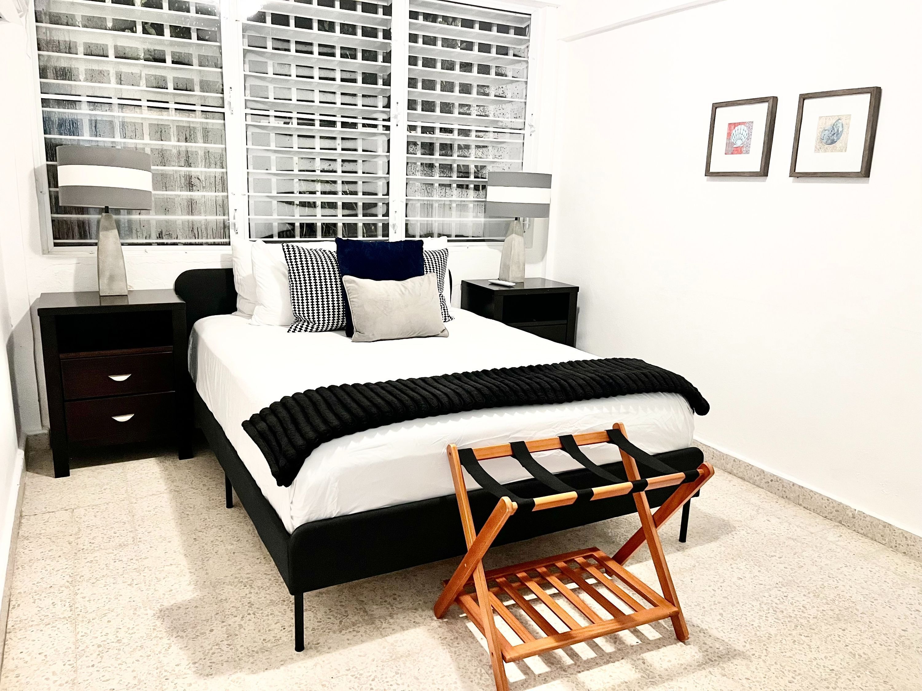 A modern bedroom with a black and white bed, a wooden chair, a nightstand, and a large window with blinds.