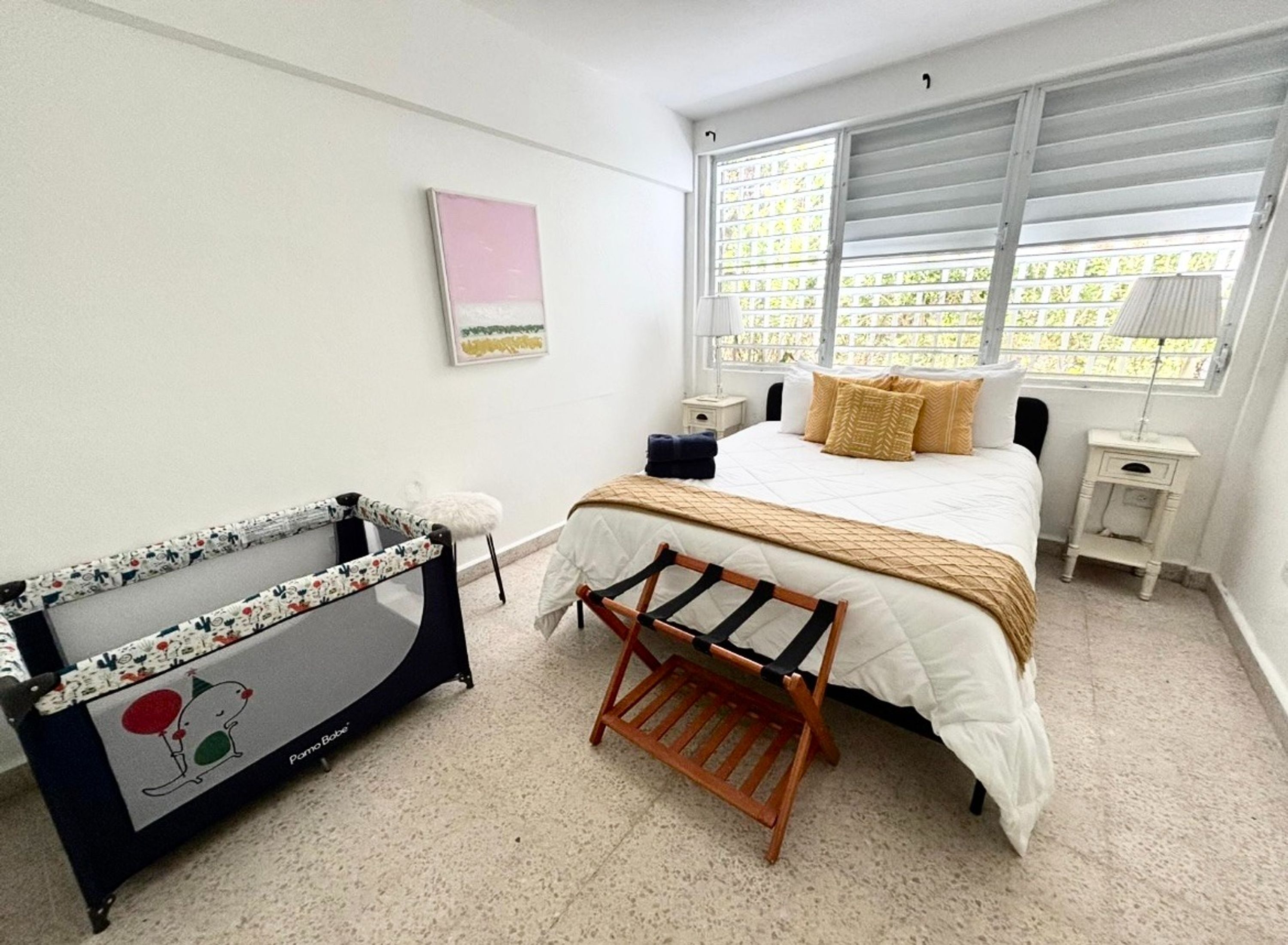 Ein Schlafzimmer mit Bett, Nachttisch, Verdunklungsvorhängen, Babybett und Wandbild.