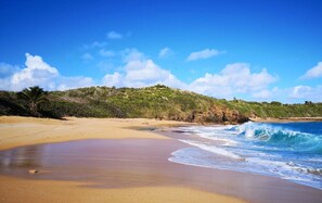 A beach with a sandy shore, waves crashing onto the beach, and a hill with vegetation in the background. - Concierge Services Walk to Beaches Restaurants (San Juan)