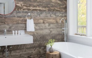 A bathroom with a wooden wall, a white sink, a mirror, a towel rack, a vase with a plant, and a window. - Stay Steps from It All – Trails, Restaurants & Shops from This Cozy Lodge (Stowe)