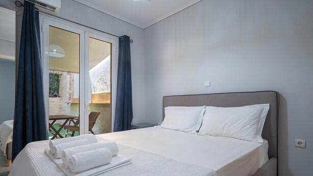 Una cama bien hecha con ropa de cama y toallas blancas, un cabecero gris y vistas a un espacio exterior a través de una puerta corredera de vidrio.