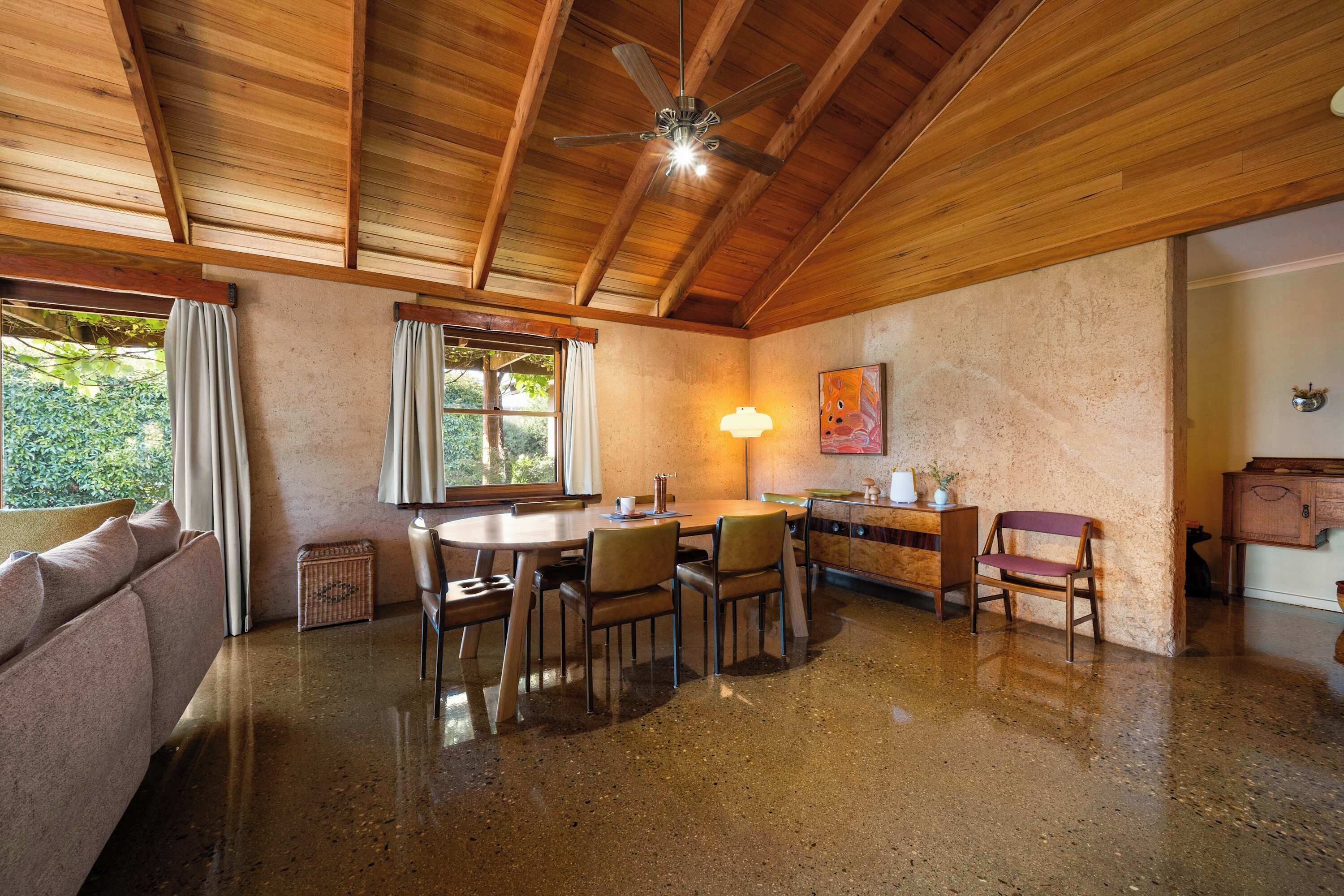 A wooden ceiling with a ceiling fan, a dining table with chairs, a sofa, a window with curtains, and a wall-mounted lamp.