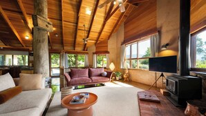 A spacious living room with a large wooden ceiling, a fireplace, a sofa, a coffee table, and a television. - Daybreak Lodge | High Country Earthen Escape (Tawonga)