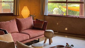 A cozy living room with a large window, a sofa, a coffee table, and a lamp. - Daybreak Lodge | High Country Earthen Escape (Tawonga)