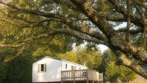 A single-story mobile home with a wooden deck, surrounded by lush greenery and trees.