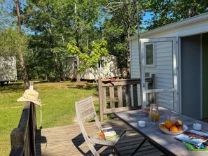 A wooden deck with a table set for breakfast, a picnic chair, and a hat on the railing. - Comfortable Mobile Home with Terrace in Charente-Maritime (Le Fouilloux)
