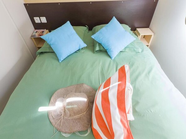 A bed with green sheets, two blue pillows, a striped orange and white pillow, and a brown hat with a bow. - Cozy mobile home with terrace in Pezuls (Pezuls)