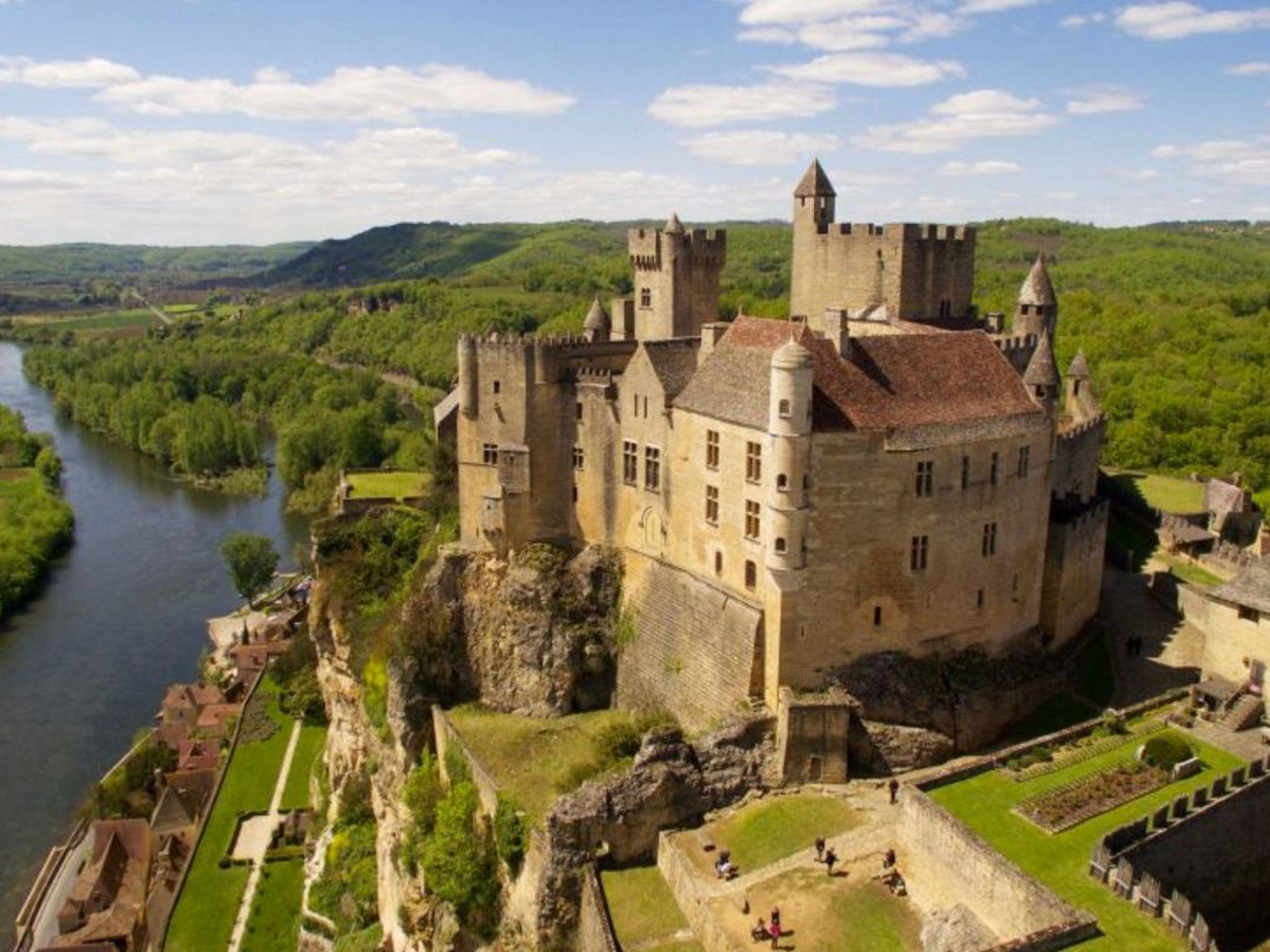 Eine Burg mit zahlreichen Türmen und einem Burggraben, umgeben von üppiger Vegetation und einem Fluss.