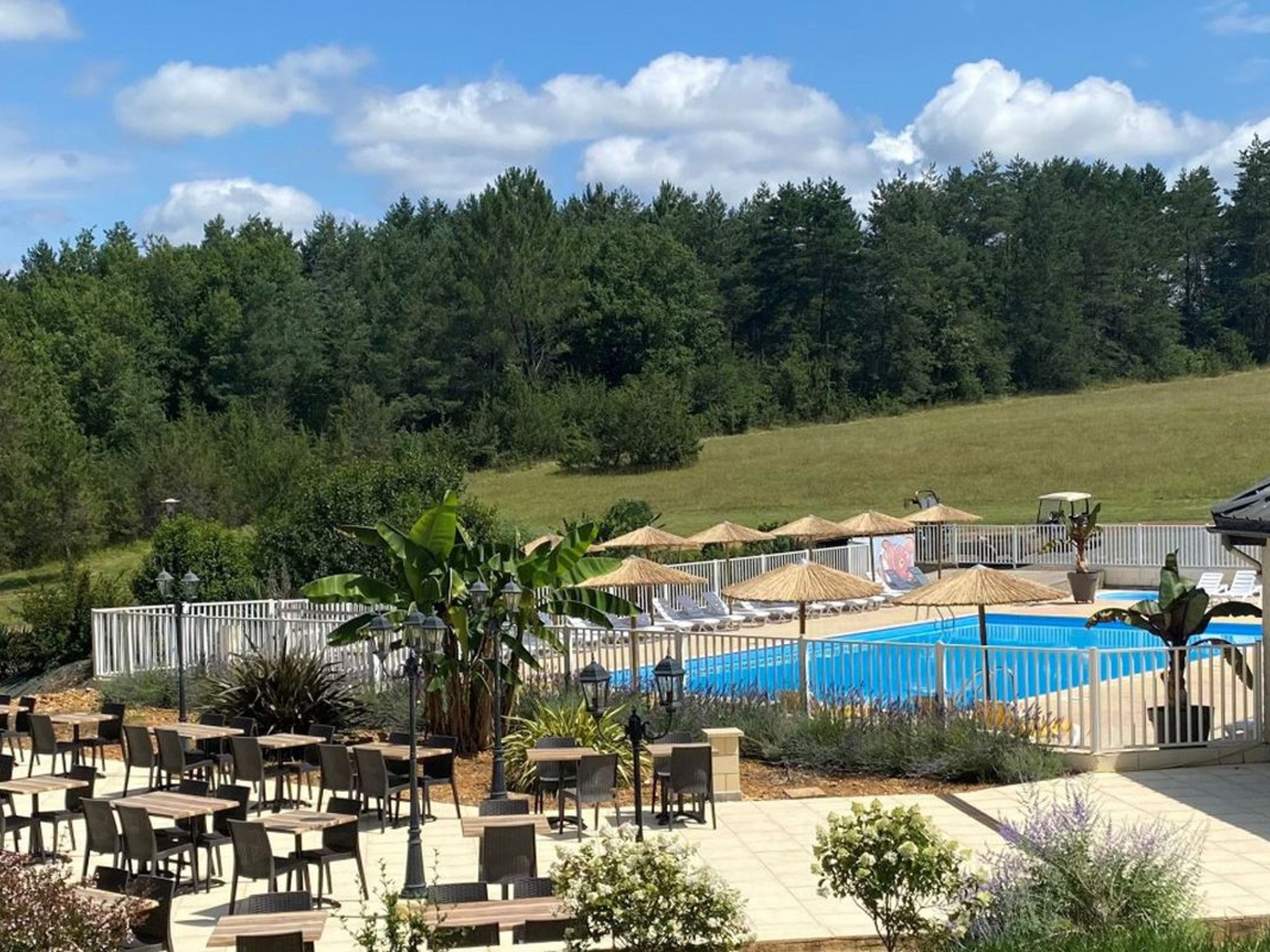 An outdoor pool area with lounge chairs and umbrellas, surrounded by a fence and greenery.