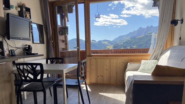 Una stanza accogliente con pavimento in legno, un tavolo bianco con sedie, un divano e una TV appesa al muro.