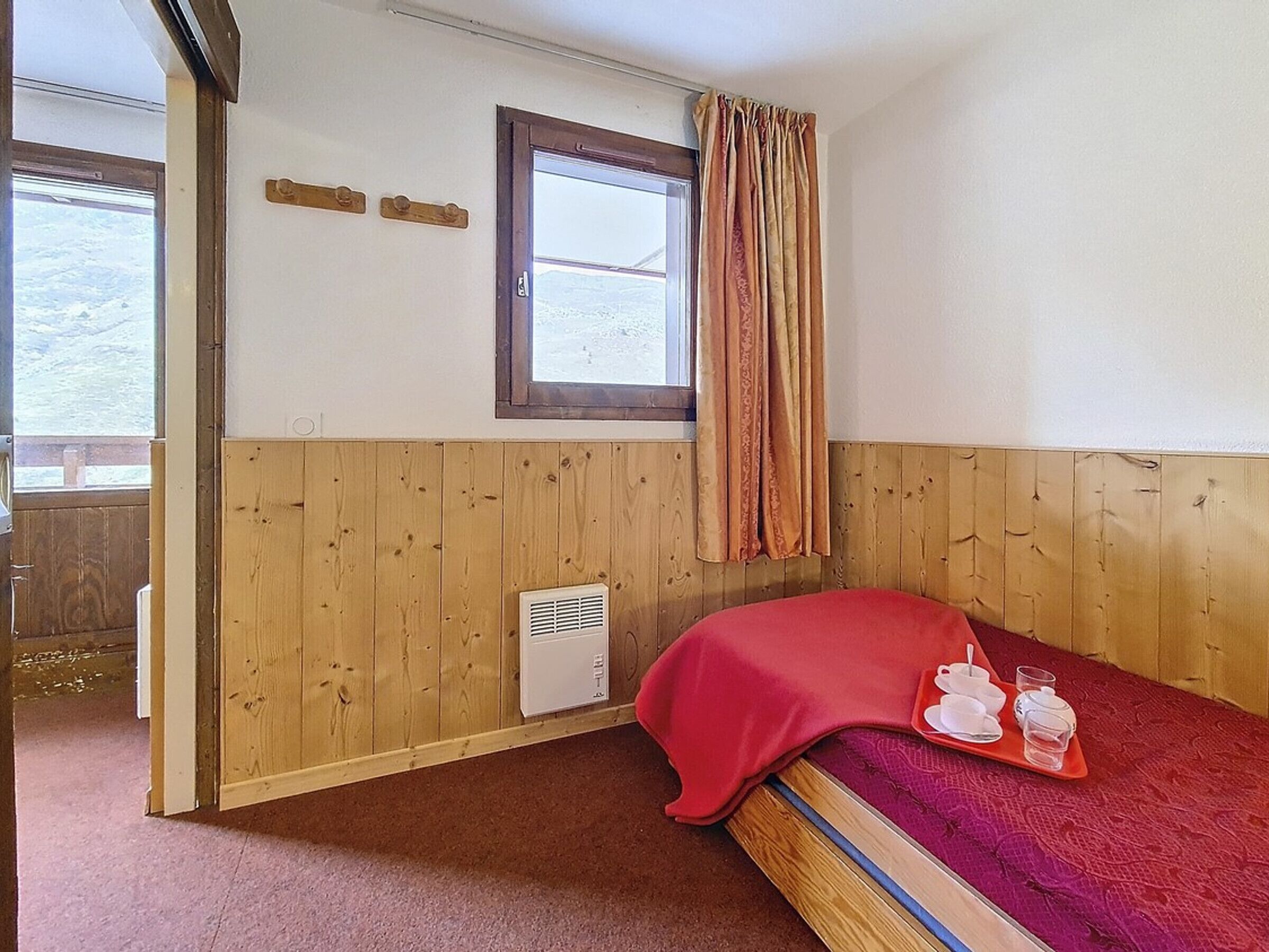 Ein Einzelbett mit roter Decke, ein Fenster mit Vorhängen, ein kleiner Heizlüfter und ein Holzwandpaneel.