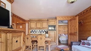 Habitación compacta con paredes de madera y suelo de baldosas, que incluye una pequeña cocina con una mesa y sillas, una cama con colcha blanca y un televisor sobre un mueble.