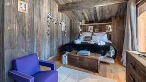 Ein rustikales Schlafzimmer mit Holzbalkendecke, einem Bett mit weißer Bettwäsche, einem violetten Sessel und einer hölzernen Kommode.