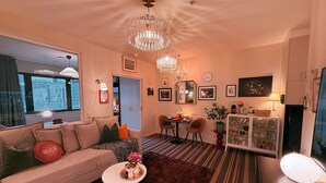 Een moderne woonkamer met een bank, salontafel en een eethoek met stoelen en een tafel. Er hangt een kroonluchter, een kast met glaswerk, en een raam met uitzicht op gebouwen.