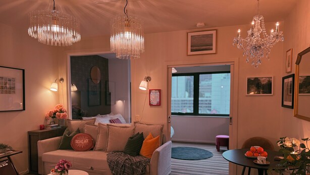 Een moderne woonkamer met een bank, een salontafel en een eethoek. Er zijn twee kroonluchters, een schilderij aan de muur en een raam met uitzicht op gebouwen.