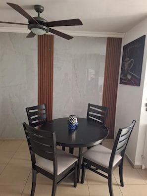 Zona pranzo con tavolo rotondo, quattro sedie, ventilatore a soffitto e un quadro appeso al muro.