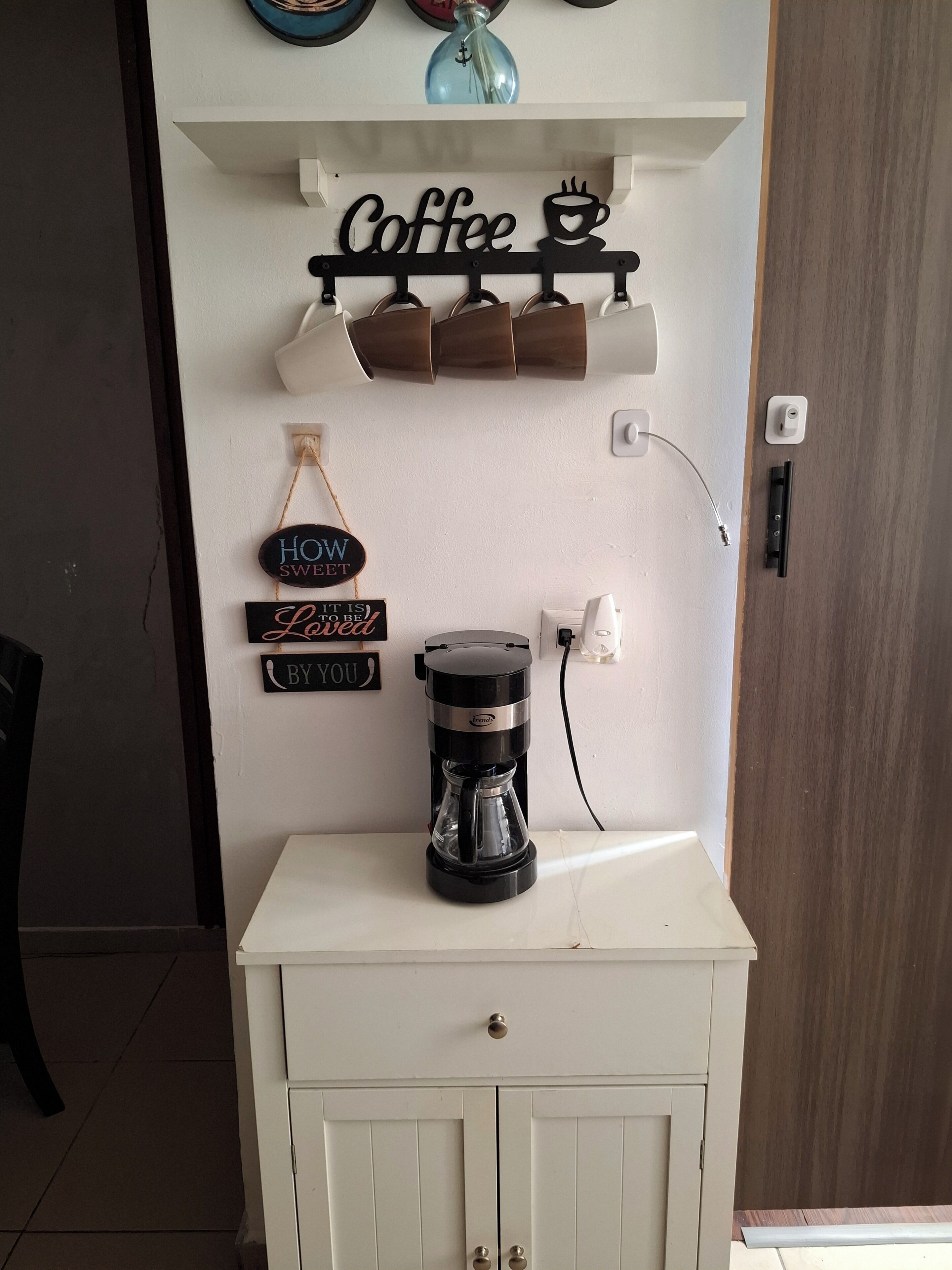 Eine Kaffeemaschine auf einem weißen Schrank, ein Wandregal mit Kaffeetassen und ein Schild mit der Aufschrift 'Coffee'.