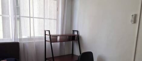 Un dormitorio con cama, un escritorio con silla, una mesita de noche de madera y una ventana con cortinas traslúcidas.
