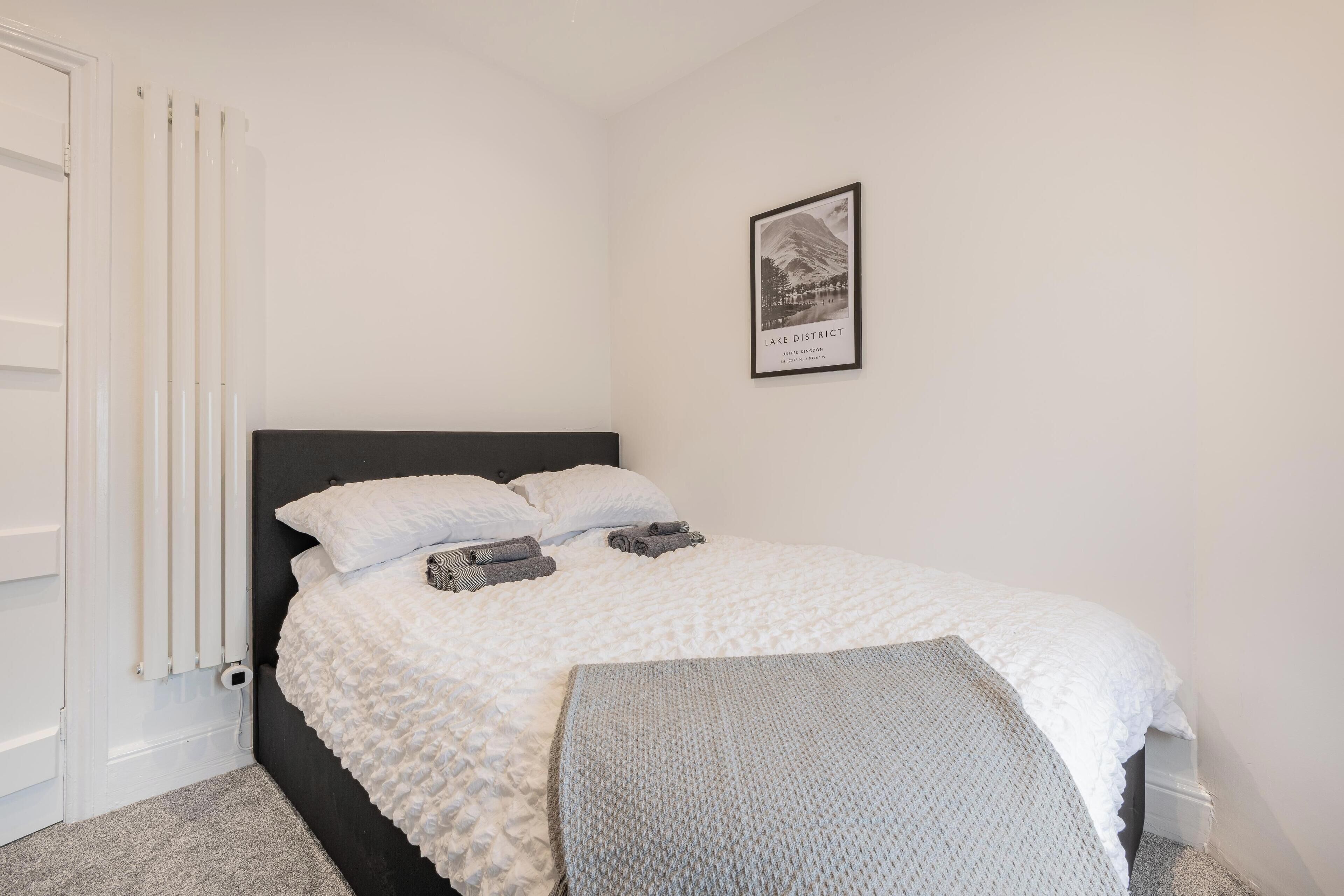 A bedroom with a bed, a radiator, a framed picture on the wall, and a grey bench.