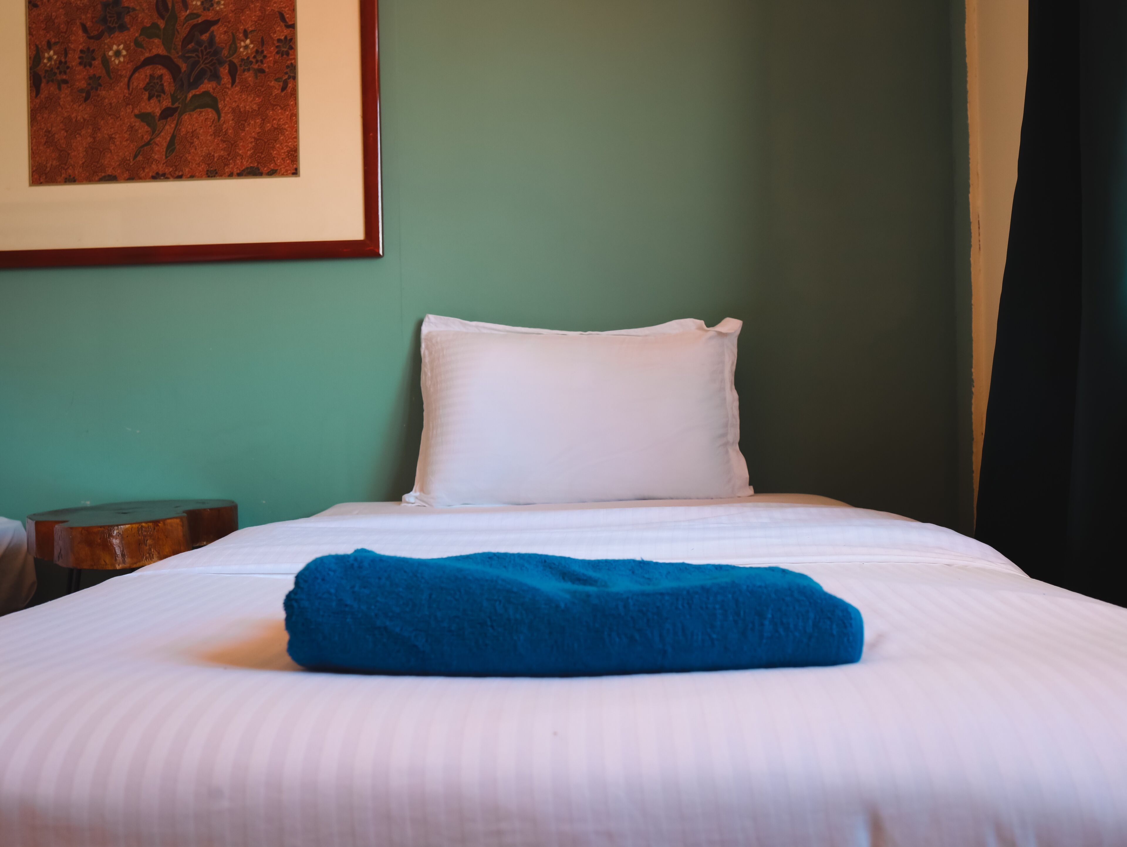 A neatly made bed with white sheets and a blue blanket, a white pillow, and a framed artwork on the wall.