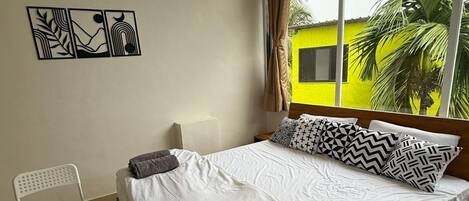 A modern bedroom with a large bed, a desk, and a chair. There is a window with a view of a yellow building and palm trees.