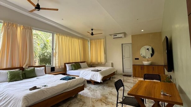 A modern hotel room with two single beds, a wooden dining table, a ceiling fan, and a bathroom with a mirror and sink. - The Tranquo Stays (Alibag)